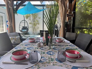 Een buiten eethoek met een tafel voor twee personen, omringd door rieten stoelen, een hangstoel en een blauw zonnescherm.