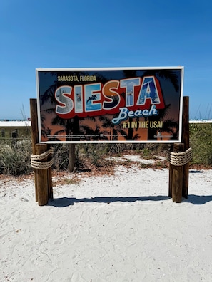 Schild für Siesta Beach in Sarasota, Florida, mit blauem Himmel und Sandstrand im Hintergrund.