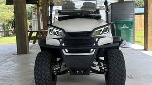 A white utility vehicle with a raised front and large tires parked under a covered area with a metal roof. - Hollywood House - Luxurious New Build With 6 Seat Golf Cart! (Bay St. Louis)