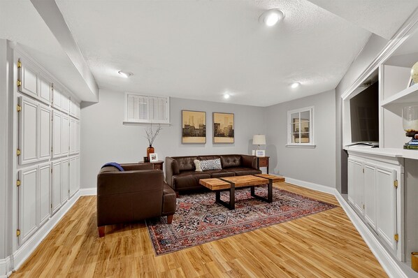 A living room with a brown leather sofa, a coffee table, a patterned rug, and a cityscape painting on the wall. - Perfect house that’s located in the heart of Indy! (Indianapolis)