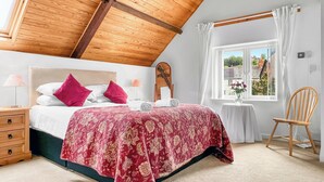 Une chambre avec un plafond en bois, un lit recouvert d’une courtepointe à motifs floraux rouges, une table de chevet avec une lampe et une fenêtre donnant sur des immeubles.