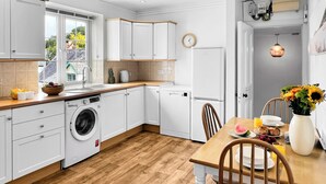 Une cuisine moderne avec des meubles blancs, un plan de travail en bois, un lave-linge encastré et un coin repas avec une table mise pour le petit-déjeuner.