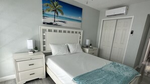 A bedroom with a bed, nightstands, a painting of a beach, an air conditioning unit, and a door. - Beach Block Apartment w/ Balcony (Clearwater)