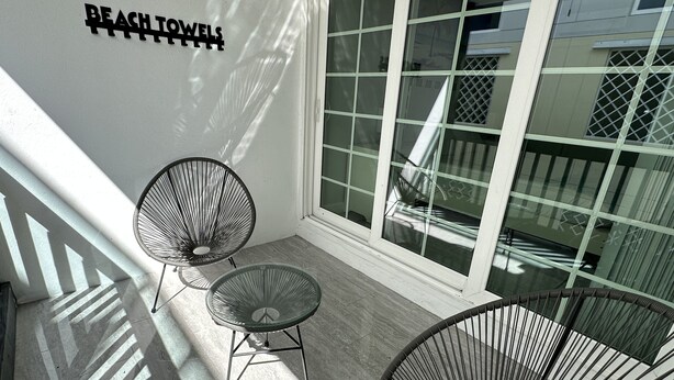 A balcony with two metal chairs and a glass table, a white wall with a sign, and a large window with a view of the outside. - Beach Block Apartment w/ Balcony (Clearwater)