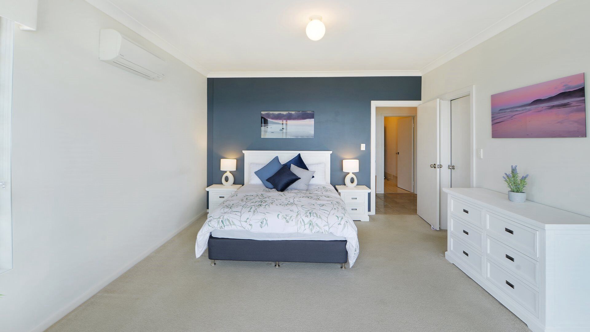 A bedroom with a bed, bedside tables, a dresser, a painting, and a hallway.