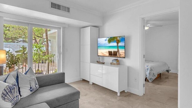 A modern living room with a grey sofa, a flat-screen TV on a white cabinet, and a view of the outdoors through large windows. - Key Biscayne Gem — Steps to Beach, Pool Views, King Bed, Parking (Key Biscayne)