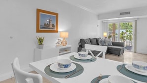 A dining area with a white table set for four, a grey sofa in the background, and a framed picture on the wall. - Key Biscayne Gem — Steps to Beach, Pool Views, King Bed, Parking (Key Biscayne)