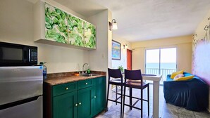 Eine kleine Küche mit Mikrowelle, Spüle und grünen Schränken. Ein Essbereich mit zwei Stühlen und einem Tisch sowie ein Balkon mit Aussicht.