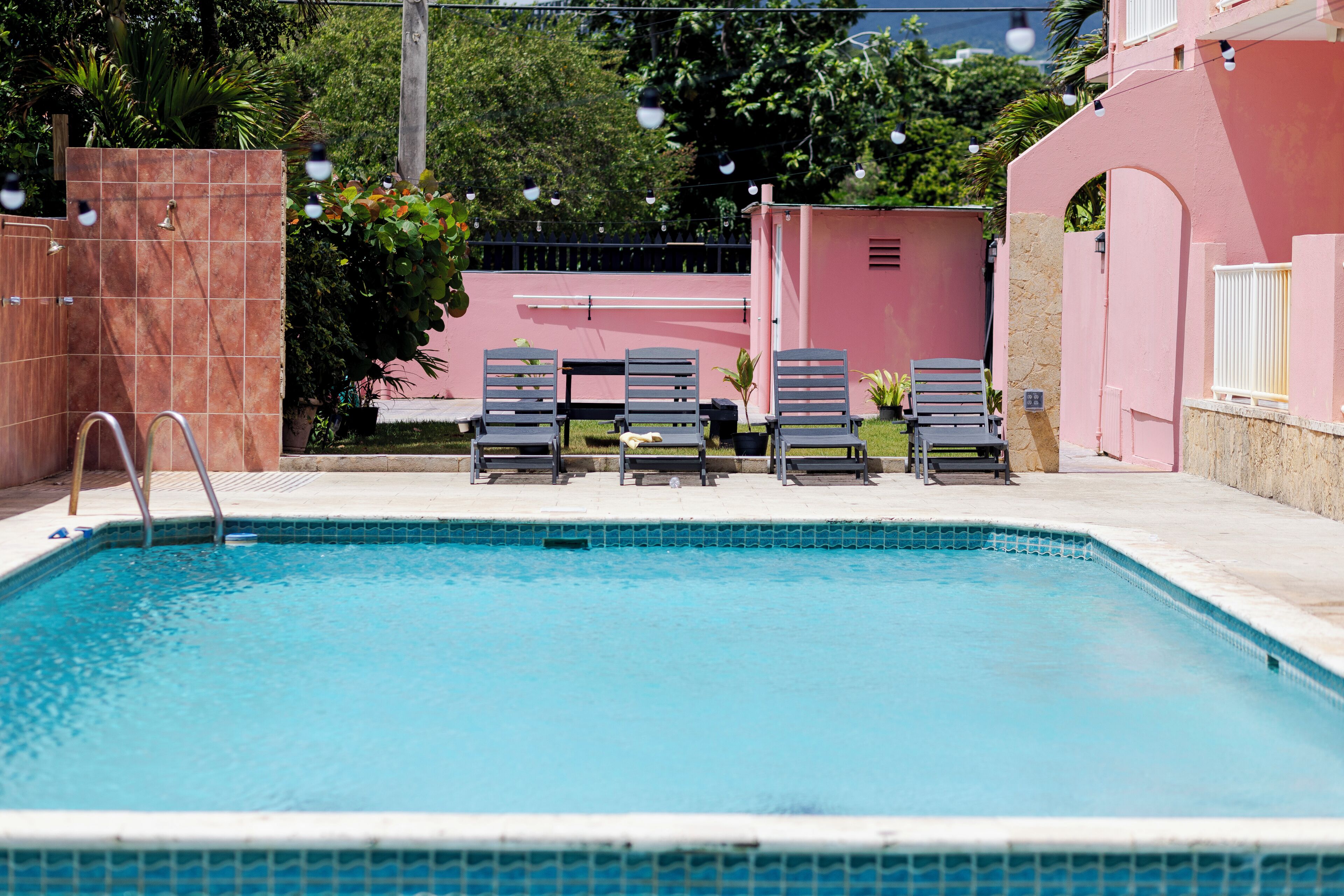 Una piscina con sillas de descanso, una terraza con baldosas y un edificio rosa al fondo.