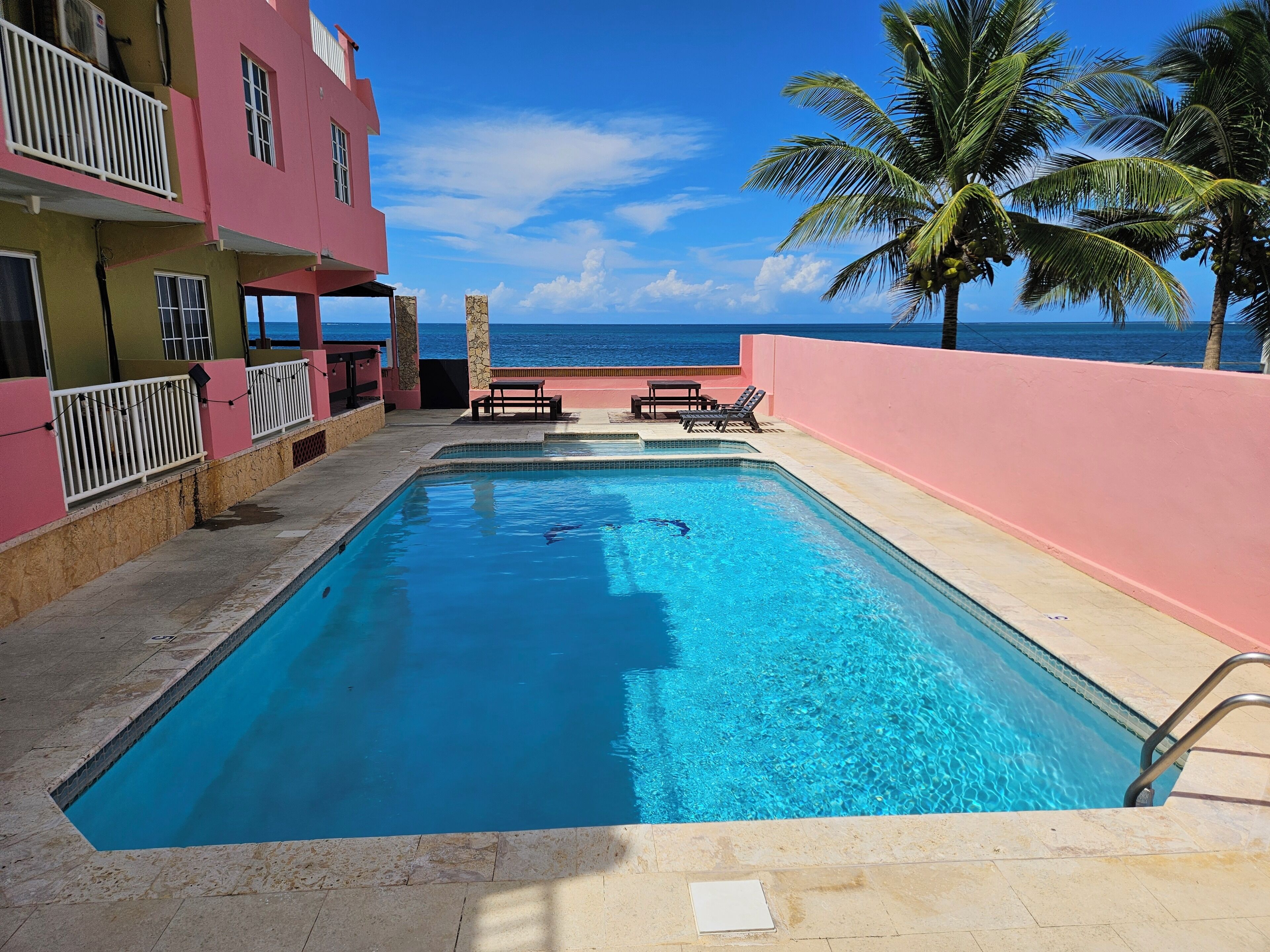 A rectangular pool with clear blue water, surrounded by a pink building with balconies and a view of the ocean.