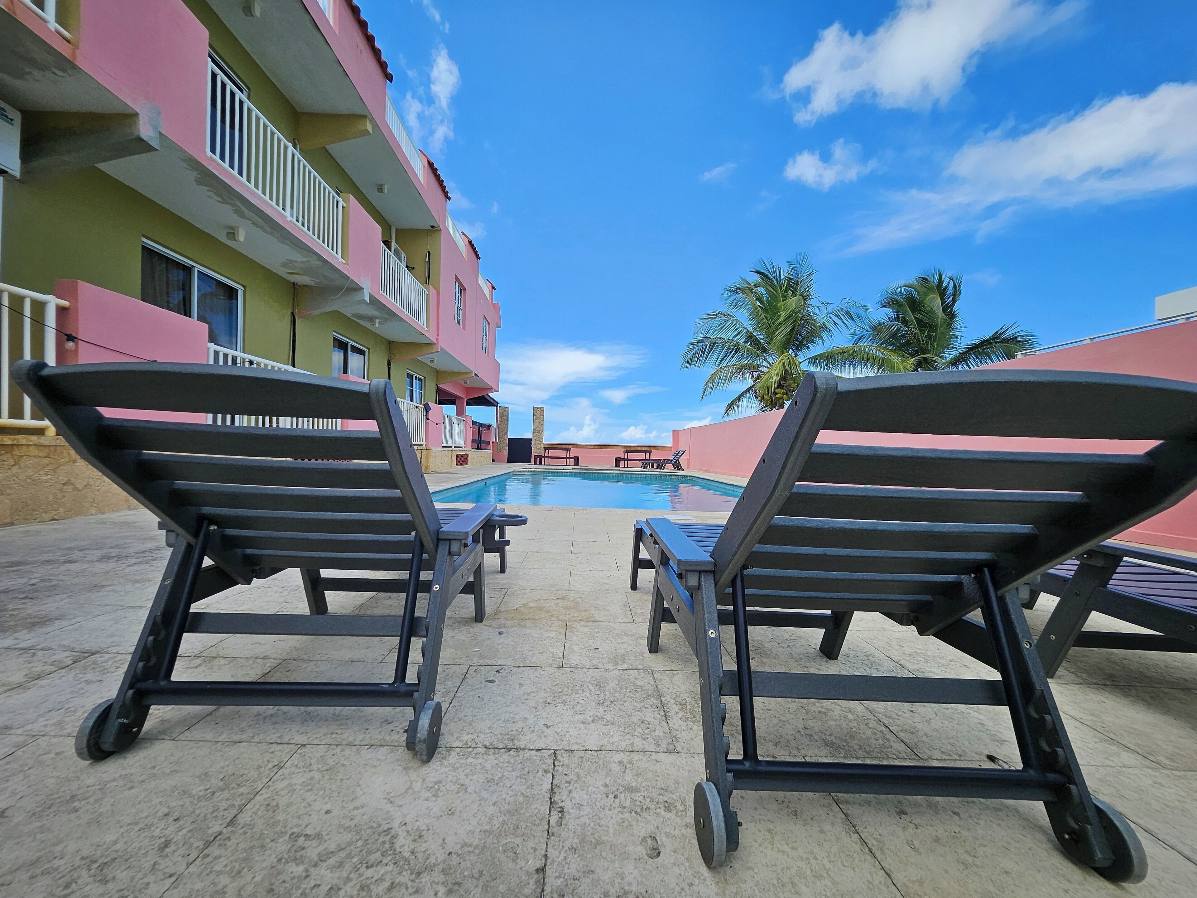 A poolside area with lounge chairs and a pool, surrounded by a building with balconies and palm trees.