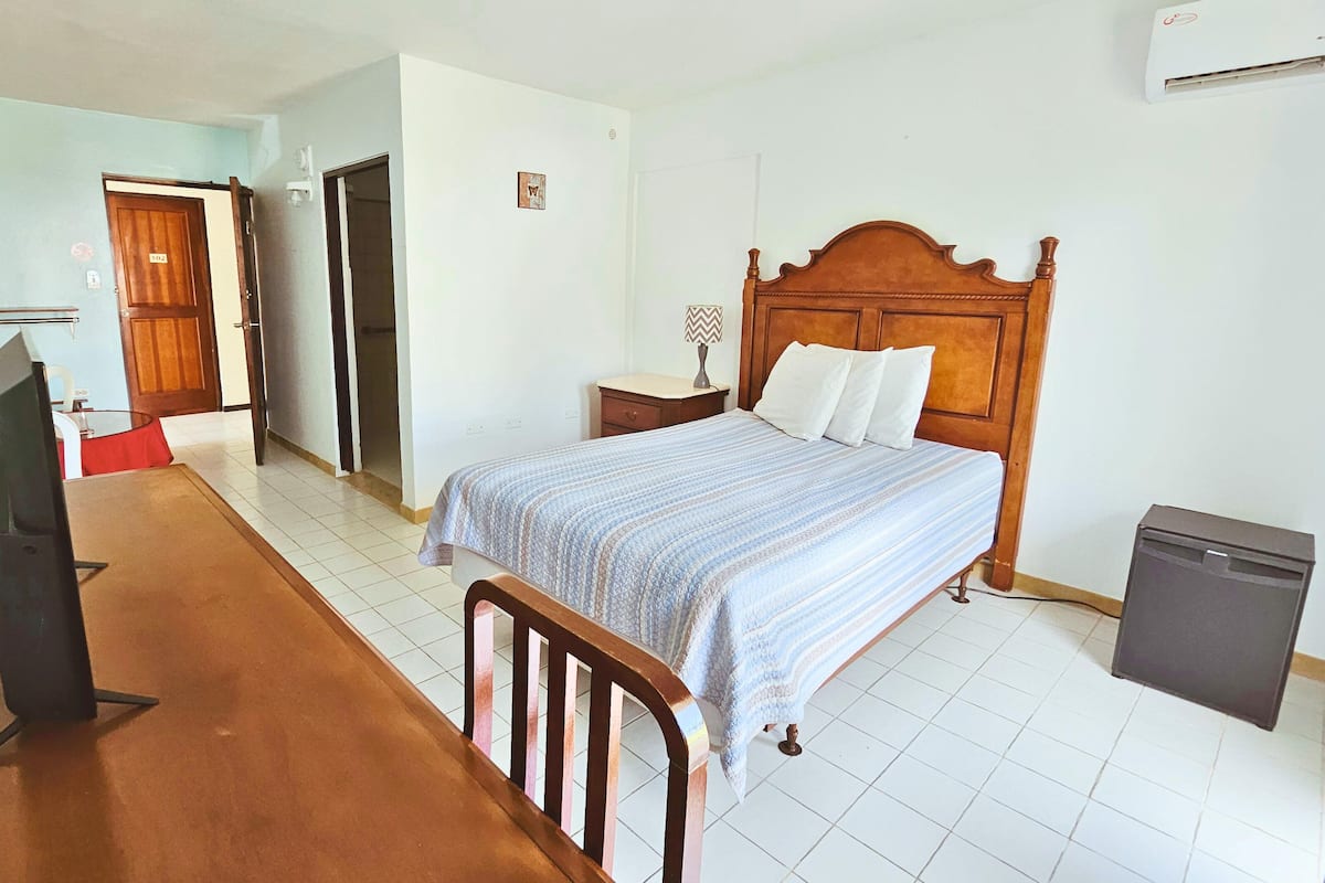 A bedroom with a bed, a desk, a chair, a nightstand, a lamp, and a small refrigerator.