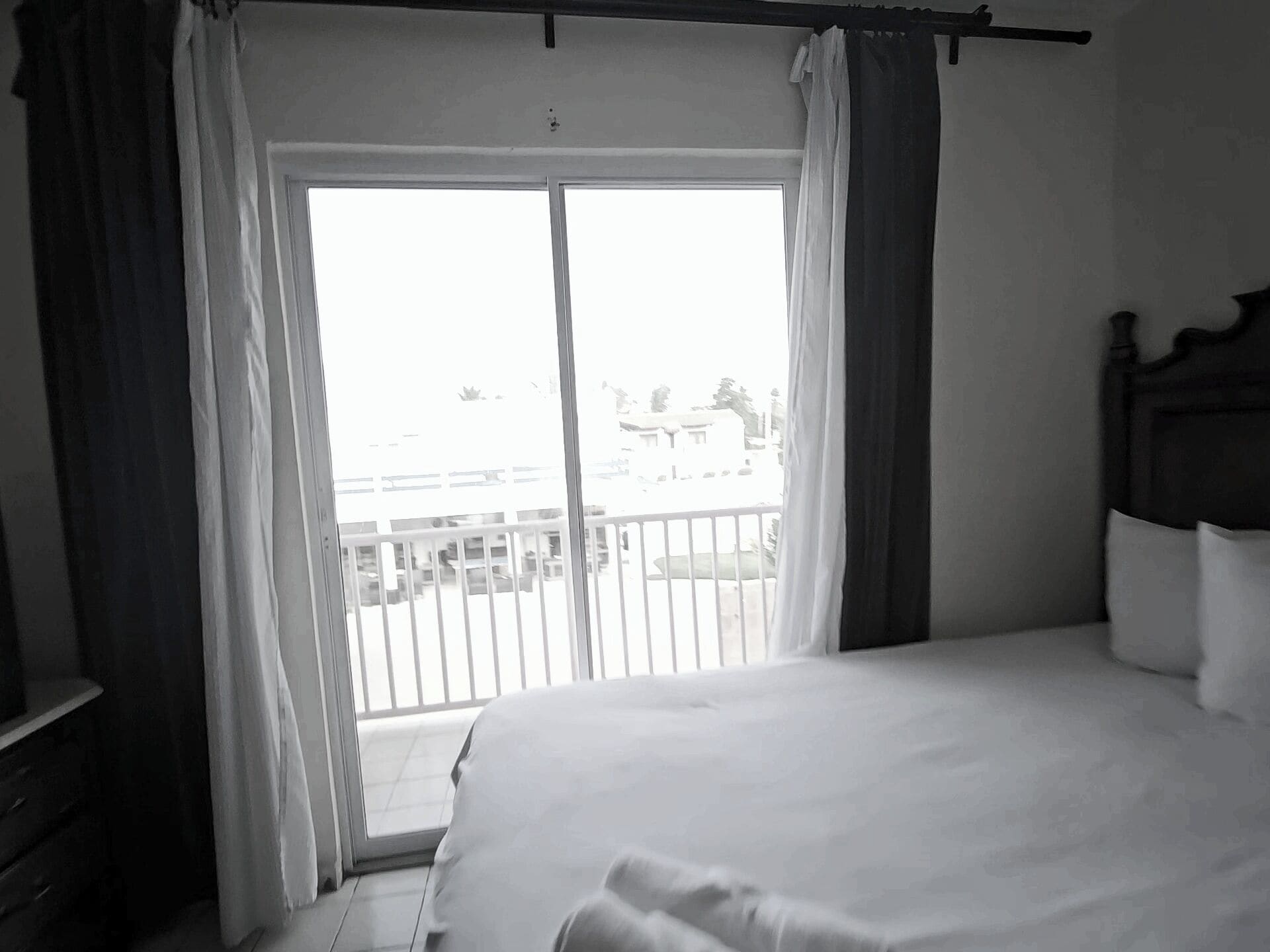 Een slaapkamer met een bed, gordijnen voor het raam, een uitzicht op een balkon en een houten hoofdeinde.
