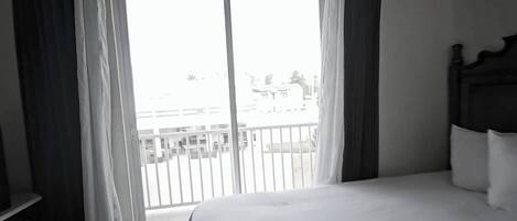 A bedroom with a bed, a window with curtains, a view of a balcony, and a wooden headboard.