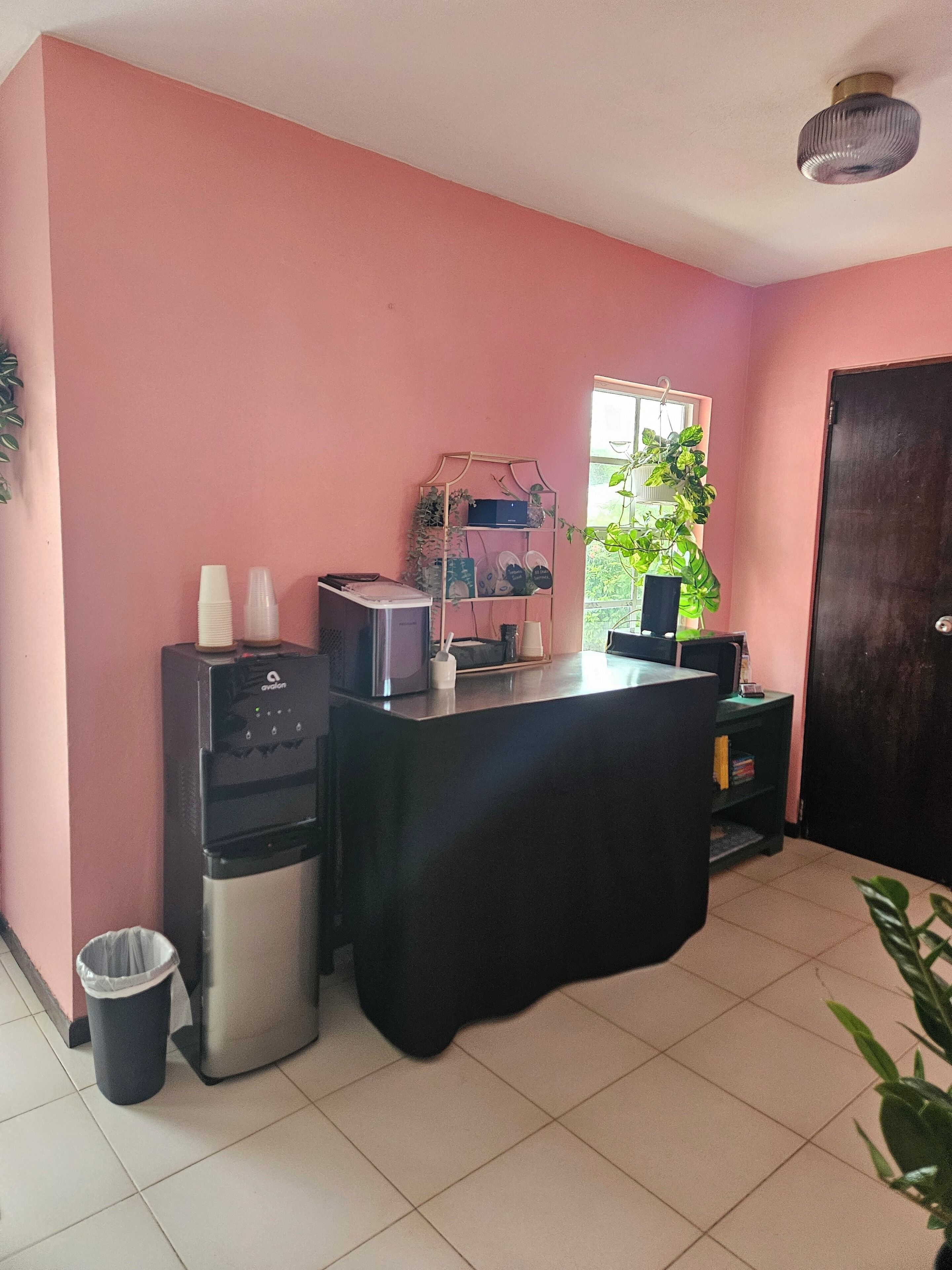Quarto com parede rosa, um bebedouro, uma máquina de café, uma pequena lixeira e uma porta.