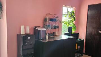 Quarto com parede rosa, um bebedouro, uma máquina de café, uma pequena lixeira e uma porta.