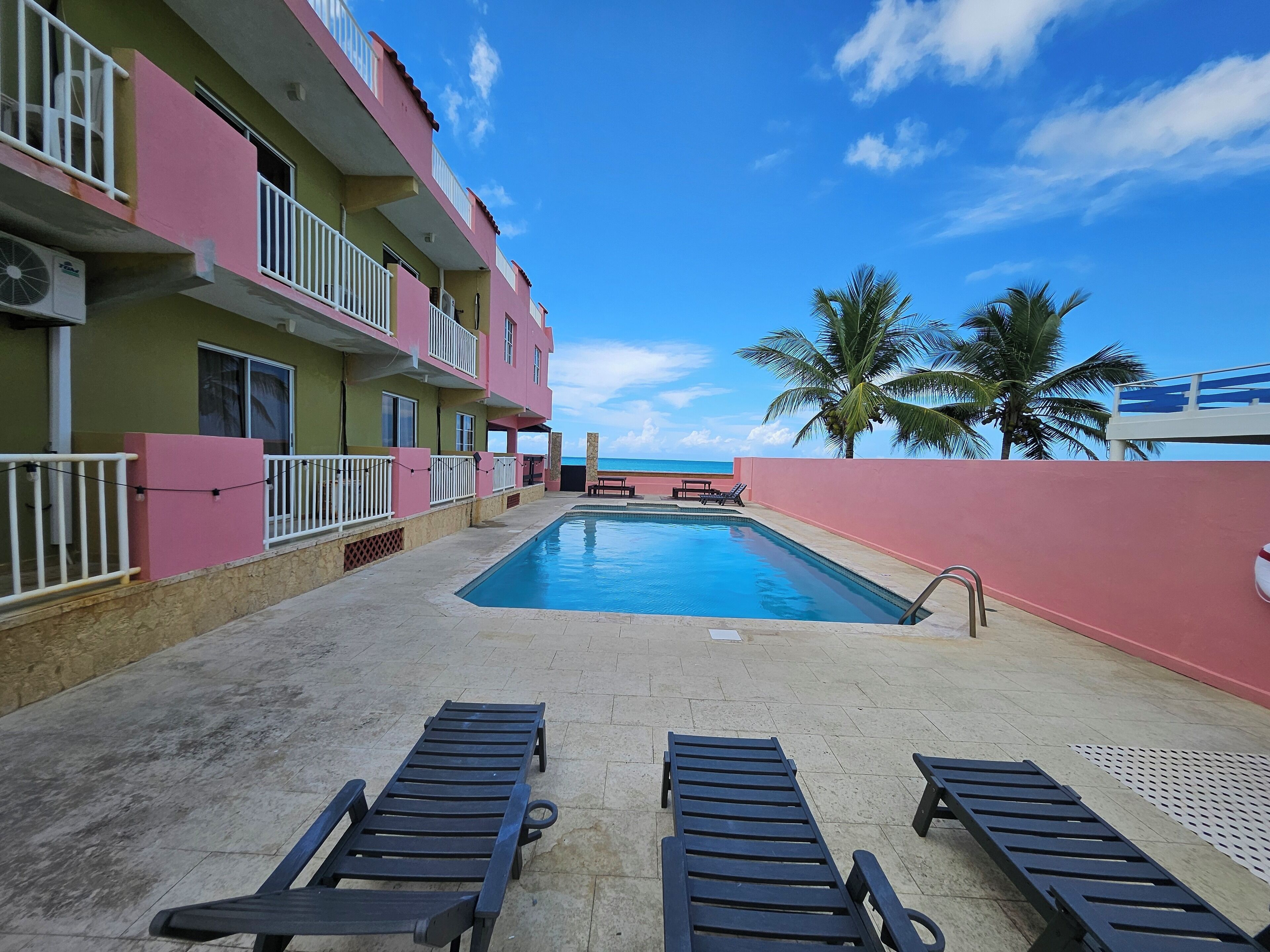 Area piscina con sedie a sdraio, vista sull'oceano e un edificio rosa con balconi.