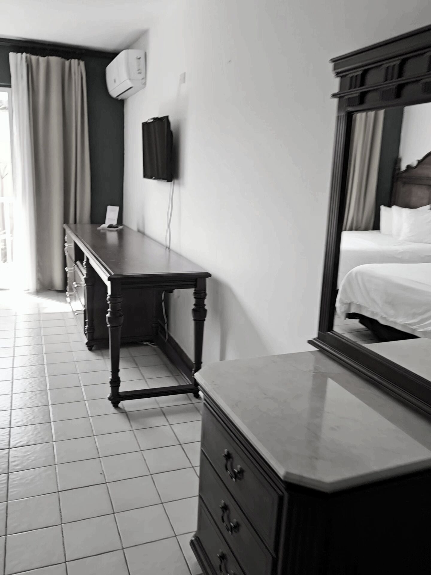 Habitación de hotel con cama, cómoda, escritorio, espejo, aire acondicionado montado en la pared y ventana con cortinas.