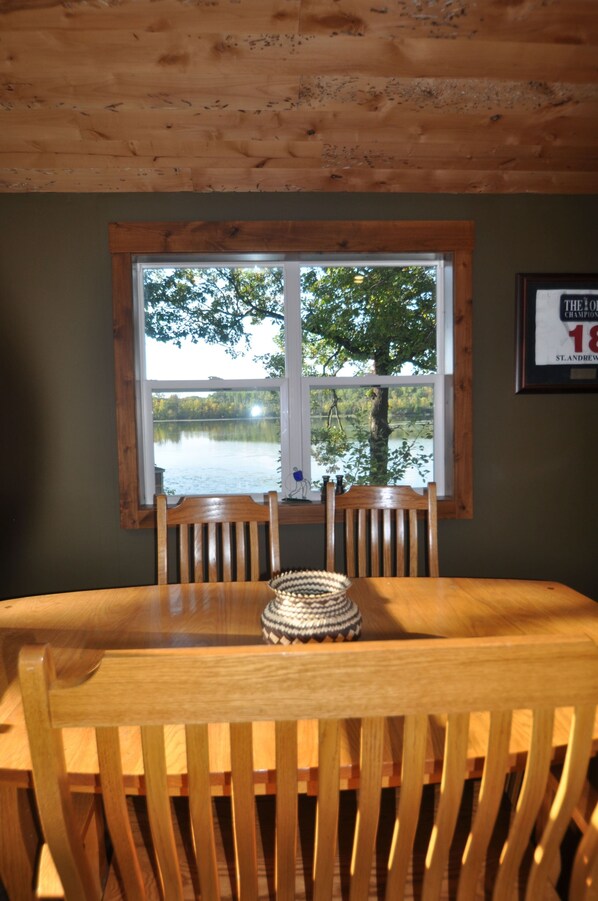 A wooden dining table with four chairs, a woven basket, and a window with a view of a lake and trees.