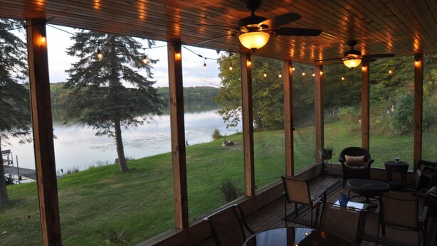 A covered outdoor seating area with wooden beams, string lights, and a view of a lake and trees. - Cuyuna Lake cabin retreat. Fish, hike, bike trails, star gazing, mins to town.  (Ironton)