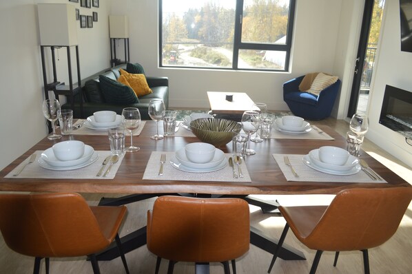 A dining area with a wooden table set for four, surrounded by orange chairs, with a view of trees and a building outside the window.