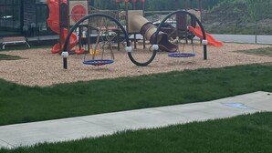 A playground with a slide, swings, and a climbing structure, set against a backdrop of mountains and a building. - Mountain View Condo w/ Hot Tube! 2B/1B - Central between Ski Resort and Downown (Revelstoke)