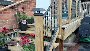 A wooden staircase with a metal railing leading to a door, surrounded by potted plants and a brick wall. - 1800s Victorian Loft  (Concord)