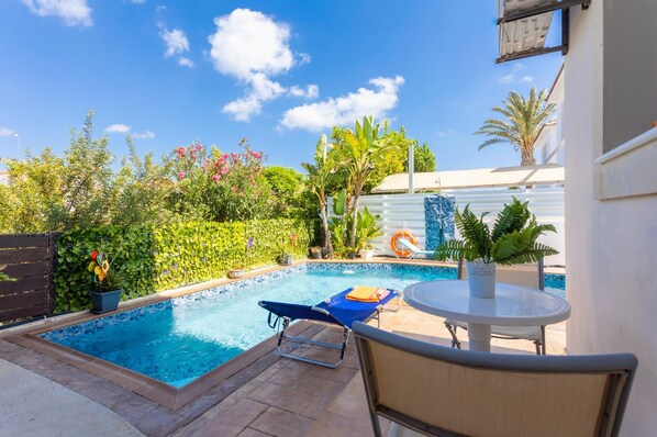 A swimming pool with a lounge chair and a table, surrounded by lush greenery and a clear blue sky.