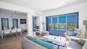 A modern living room with a sofa, coffee table, and TV. There is a dining area with chairs and a table. Large windows offer a view of the sea. - Villa Kalliopi in Pernera (Paralimni)