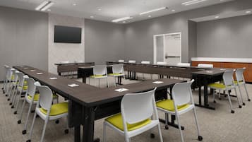 Uma sala de conferências com várias mesas e cadeiras, uma televisão de tela grande na parede e uma porta ao fundo.