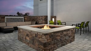 Een terras buiten met een vuurkorf van steen, groene stoelen en een witte kolom.