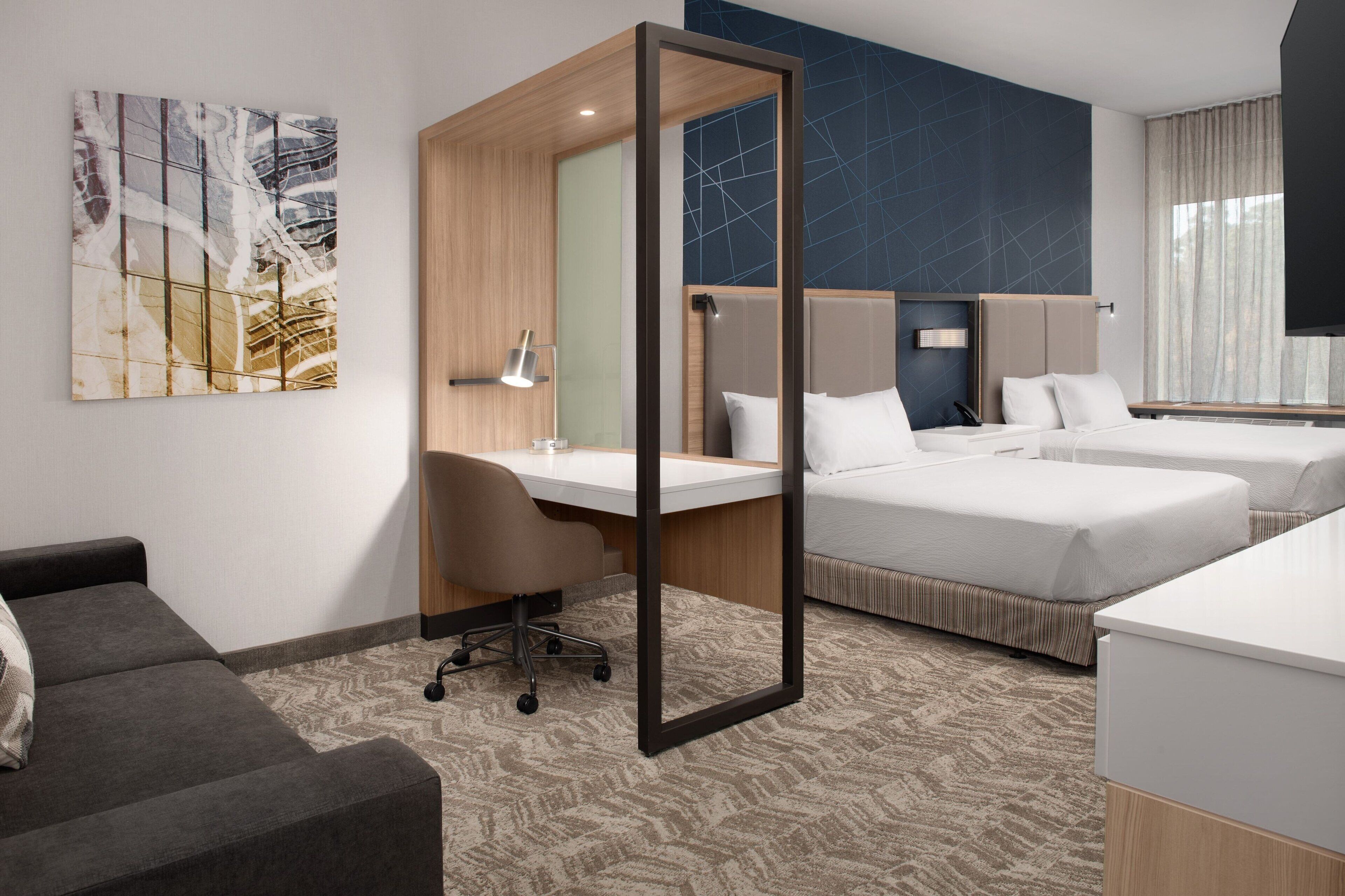 A modern hotel room with a desk, two beds, a sofa, and a large abstract painting on the wall.