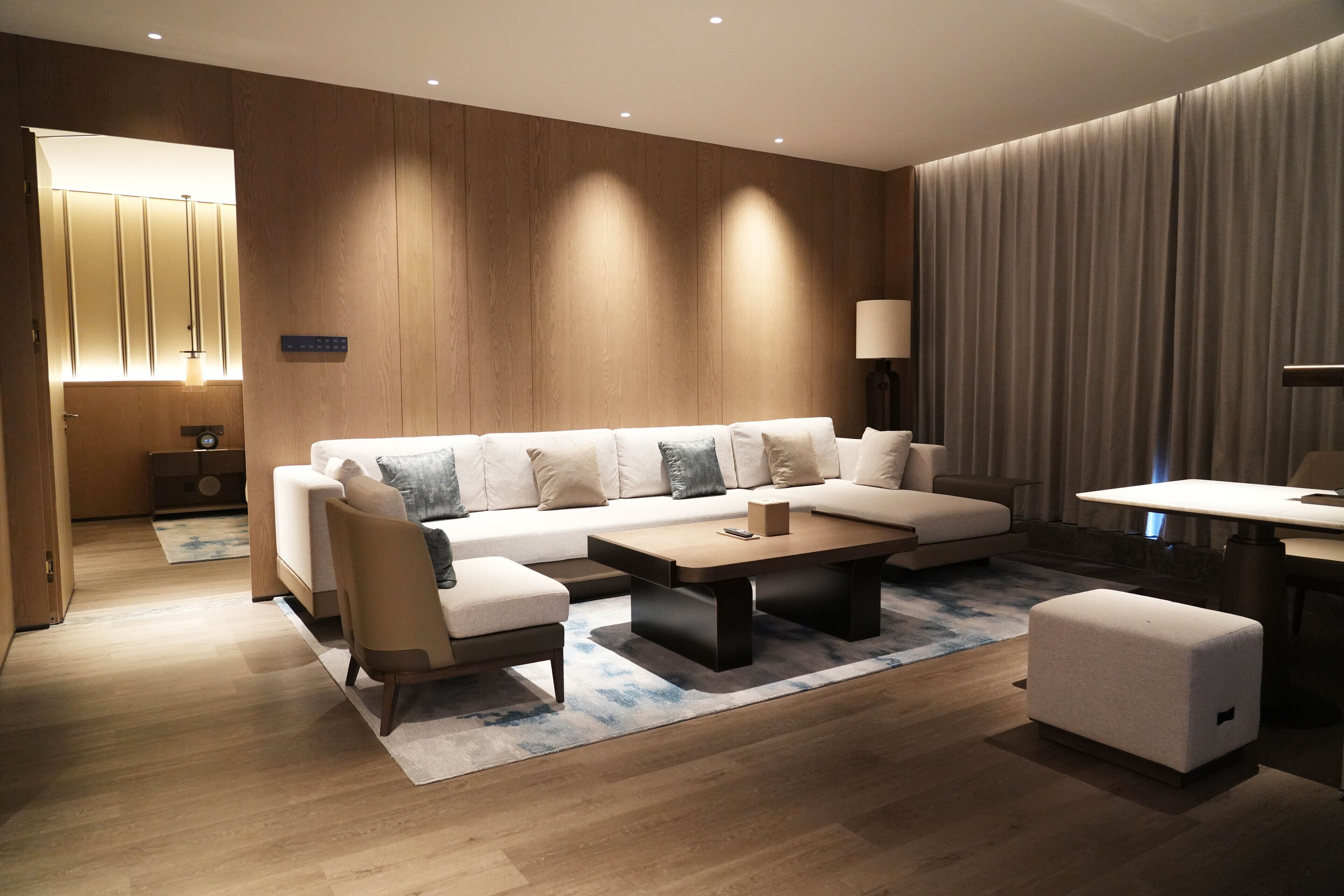 Una sala de estar moderna con un sofá, una mesa de centro y un reposapiés. Se observa una lámpara, una mesita auxiliar y un revestimiento de pared de madera.