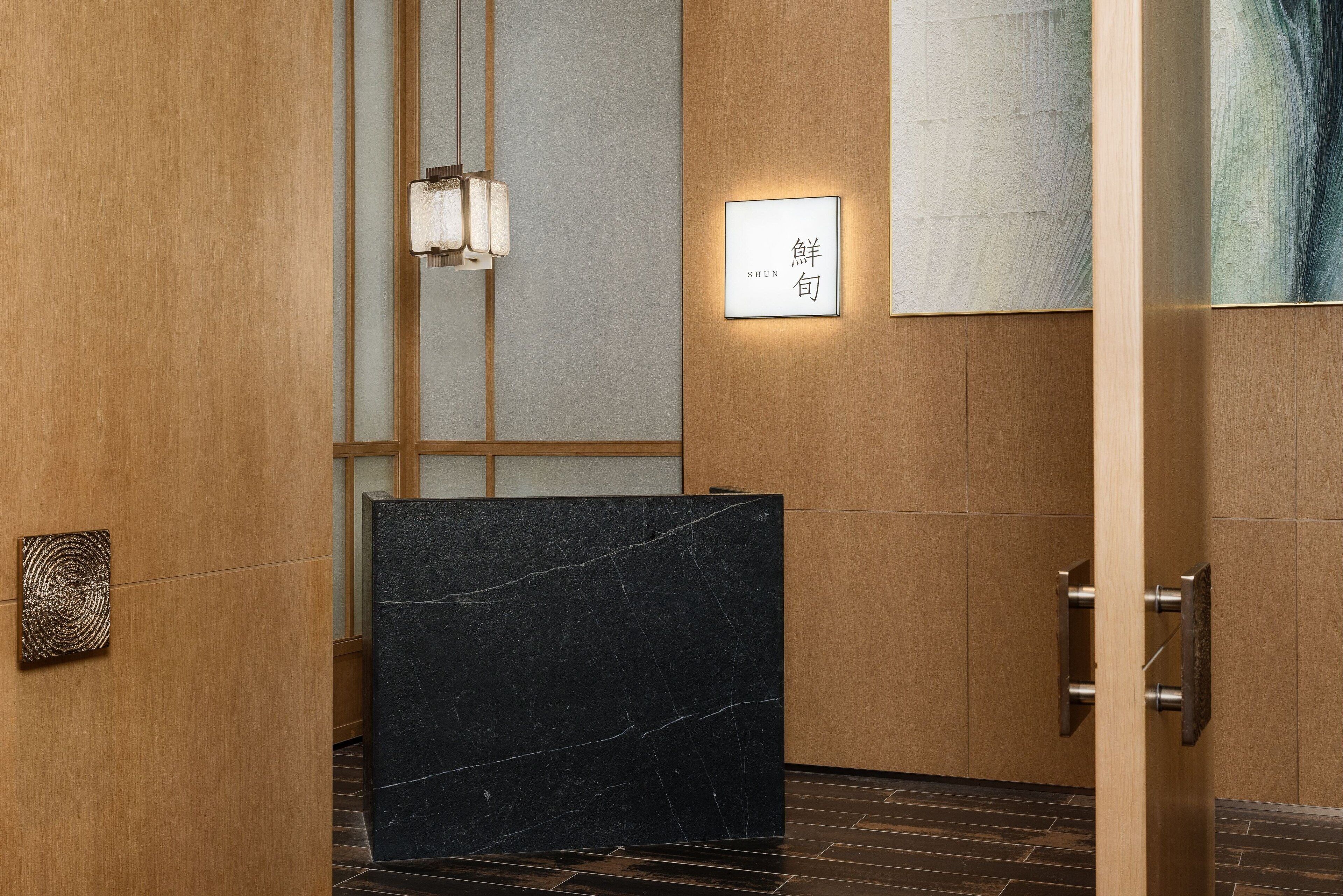 Uma área de recepção com balcão de mármore preto, painéis de madeira e luminária pendente.