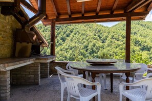 Ein überdachter Essbereich im Freien mit Kamin aus Stein, weißen Kunststoffstühlen und Blick auf einen bewaldeten Berg.