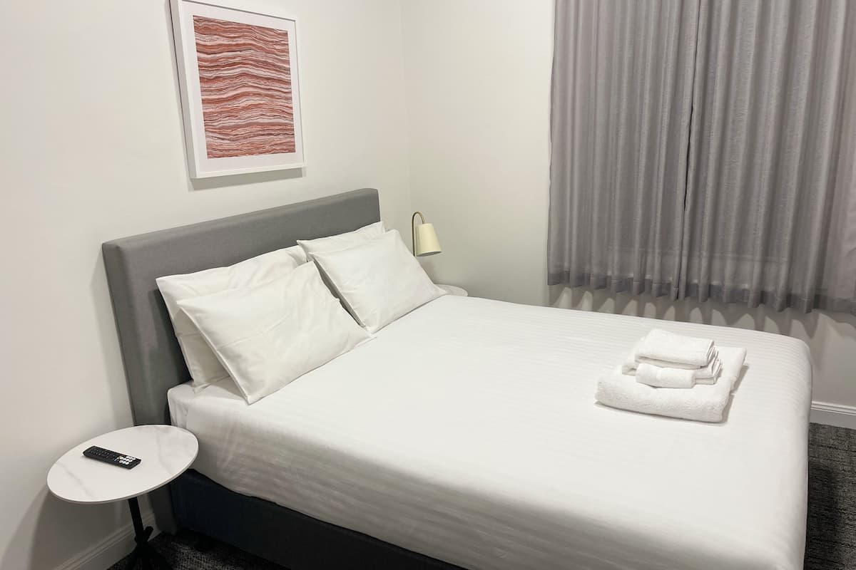A neatly made bed with white linens, a grey headboard, and a small bedside table with a remote control.