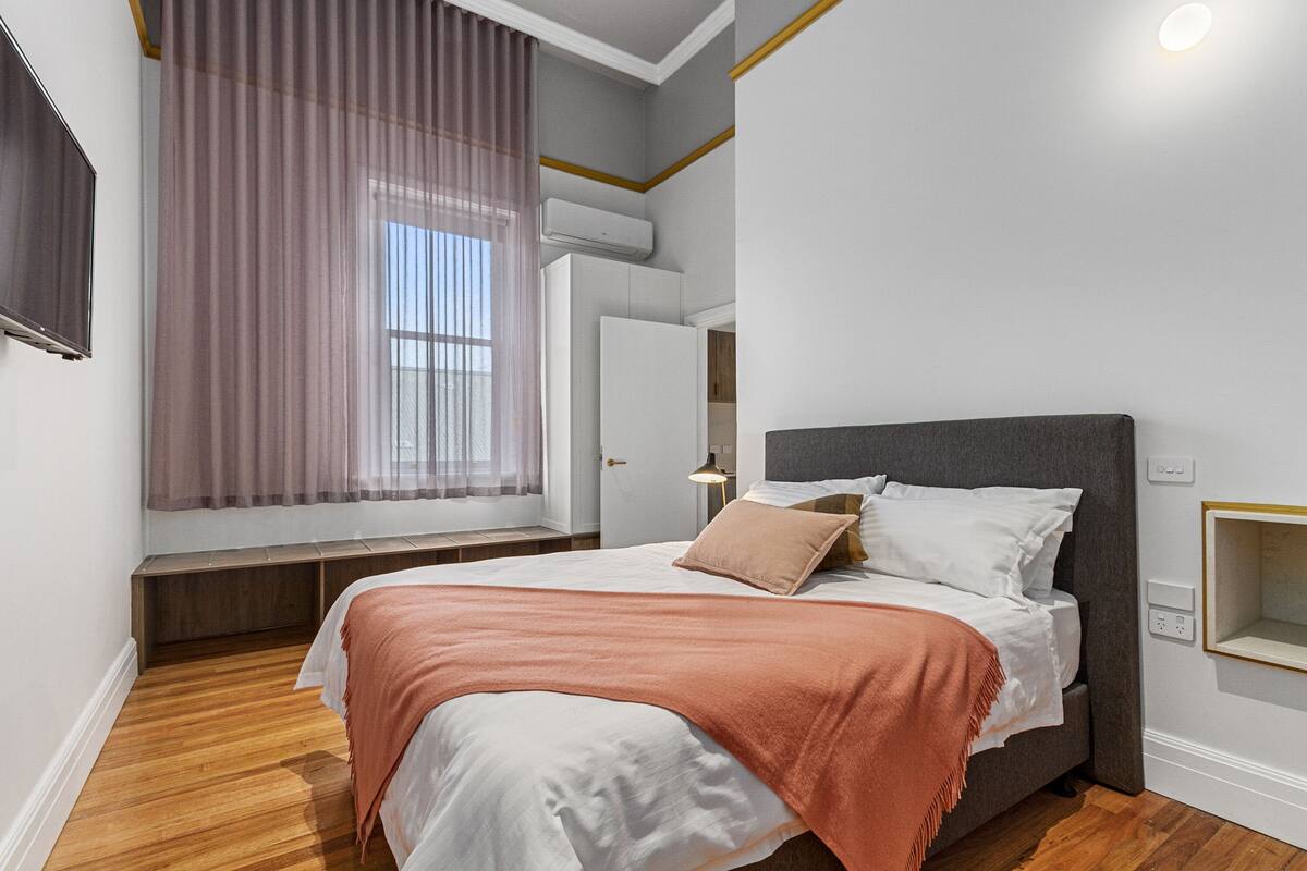 A modern bedroom with a large bed, a bench at the window, and an air conditioning unit above the door.