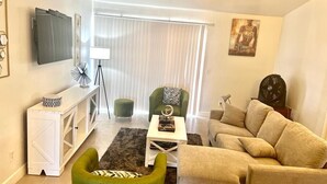 Uma sala de estar com um sofá bege, uma poltrona verde, uma mesa de consola branca e uma TV fixada na parede.