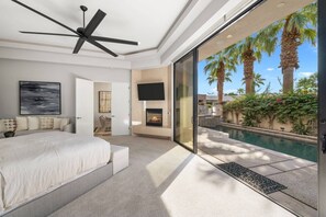 A bedroom with a large bed, a ceiling fan, a fireplace, and a view of a swimming pool with palm trees.