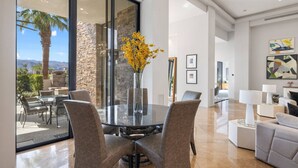 Un interior moderno con una mesa de comedor de vidrio rodeada de sillas, una pared de acento de piedra y vistas a palmeras y montañas.