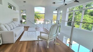Un salon spacieux avec un canapé blanc, un fauteuil blanc et une petite table. De grandes fenêtres offrent une vue sur la verdure environnante.
