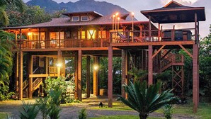 A two-story wooden house with a balcony, surrounded by lush greenery and mountains in the background. - The Iconic Bamboo House (Hanalei)