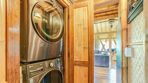A laundry area with stacked washers and dryers, wooden walls, and a view into a living area with a couch and a hanging light fixture. - The Iconic Bamboo House (Hanalei)