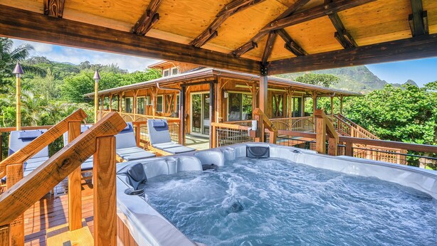 A wooden deck with a hot tub, lounge chairs, and a covered area with a view of lush greenery and mountains. - The Iconic Bamboo House (Hanalei)