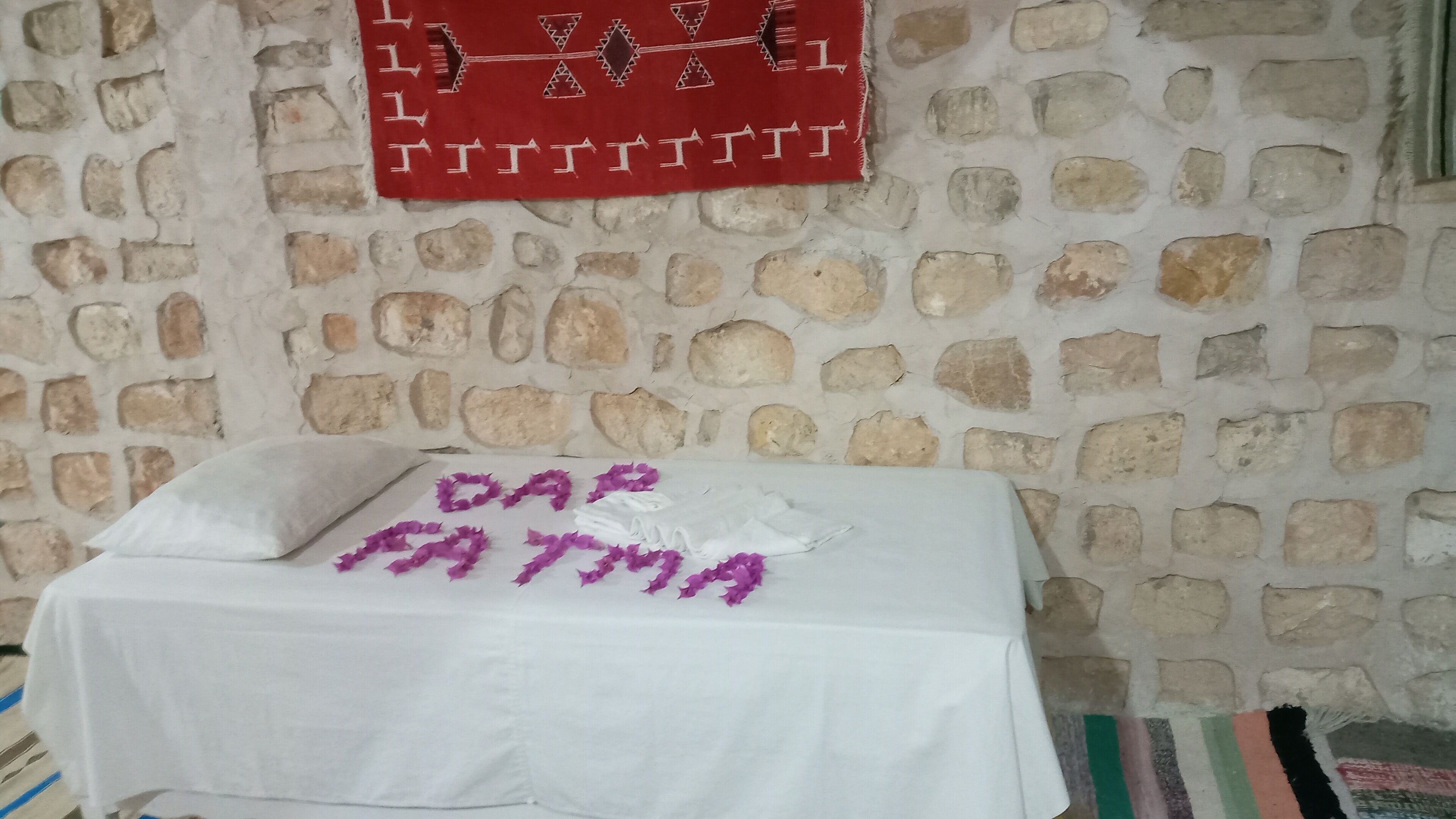Habitació amb paret de pedra, llit amb full blanc i un objecte decoratiu vermell a sobre.