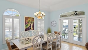 A dining area with a white table and chairs, a chandelier, a clock, and a window with shutters. - Mango Margarita- Beach Home with Pool, Sleeps 12 (Fort Myers Beach)