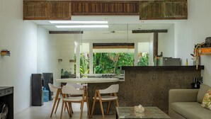 A modern living room with a sofa, coffee table, and dining area with chairs. There is a kitchen visible in the background with a refrigerator and a water dispenser. - The Saya House Villa by Betterplace (Kerobokan)