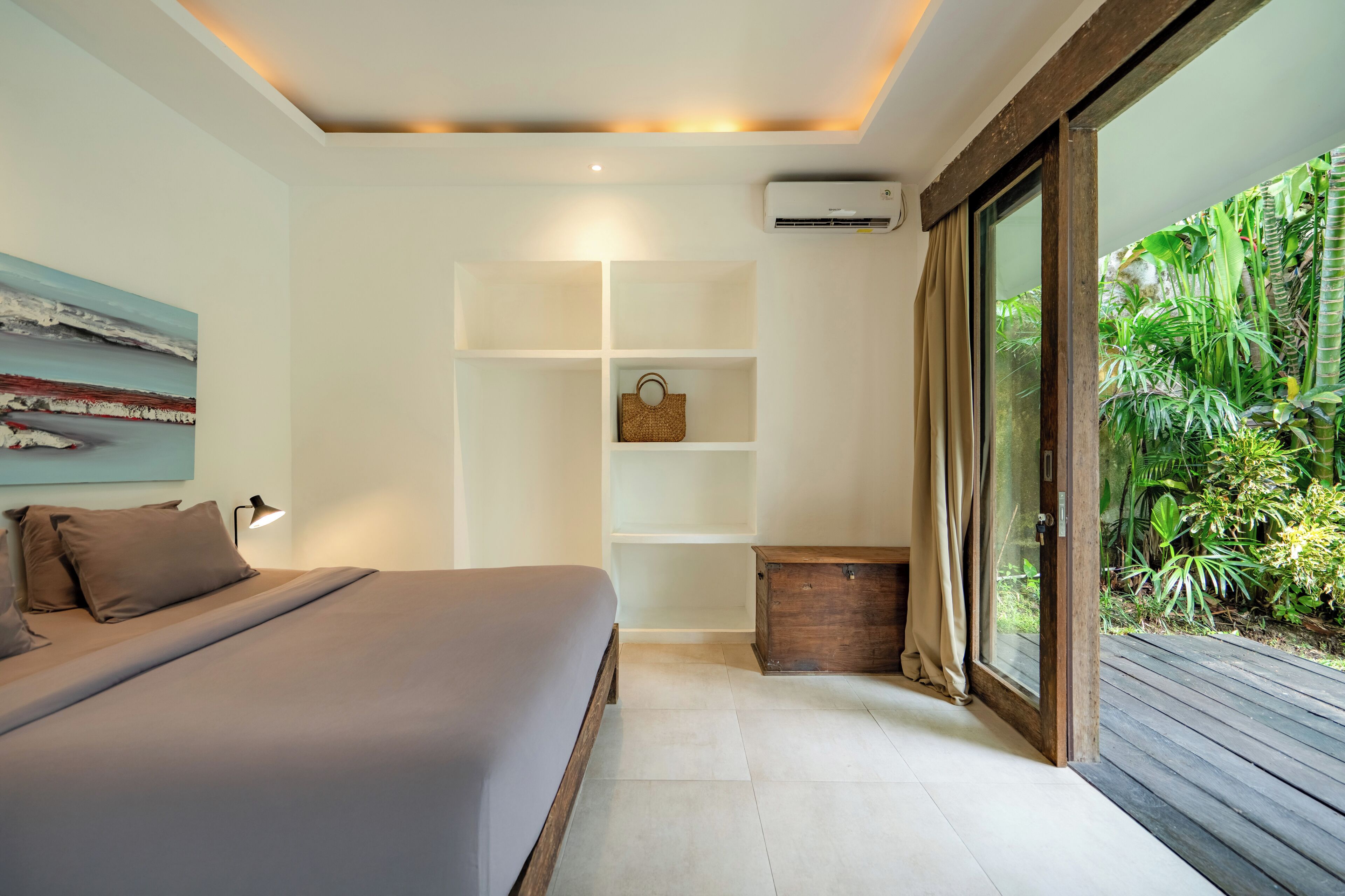 Un dormitorio con una cama, una mesita de noche, un estante en la pared, una ventana con cortinas y vistas al exterior.
