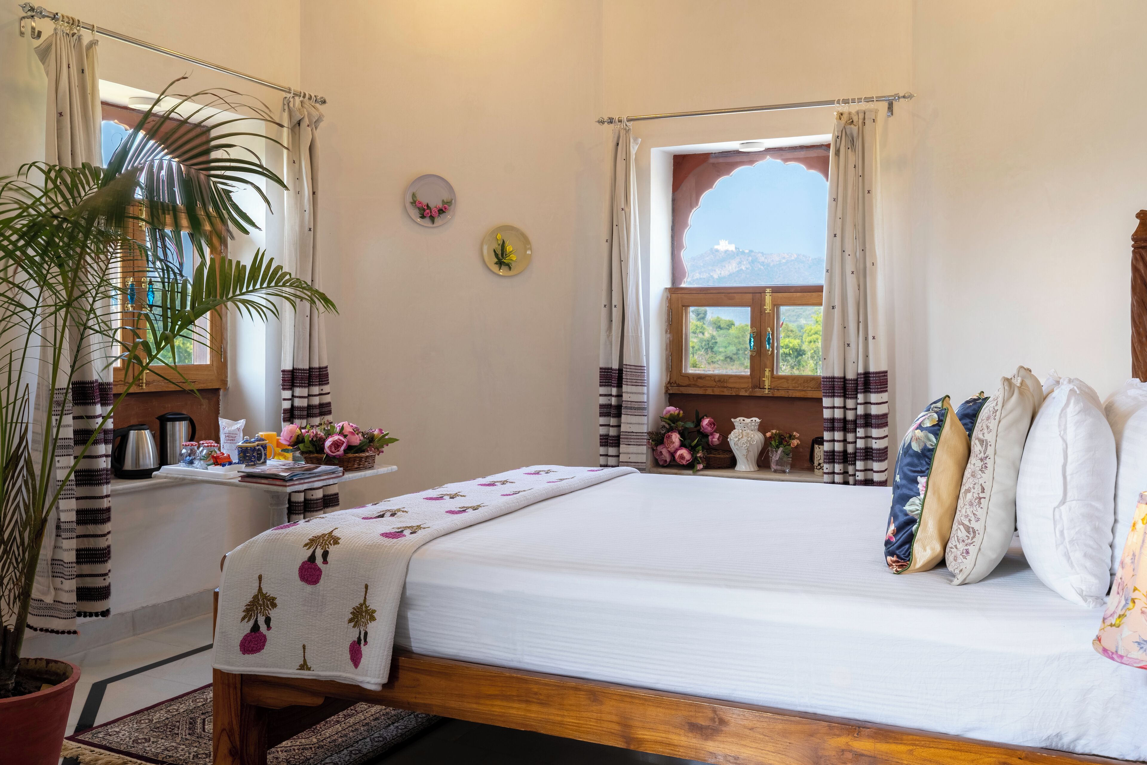 A bedroom with a large bed, a potted plant, a window with curtains, and a view of the outdoors.