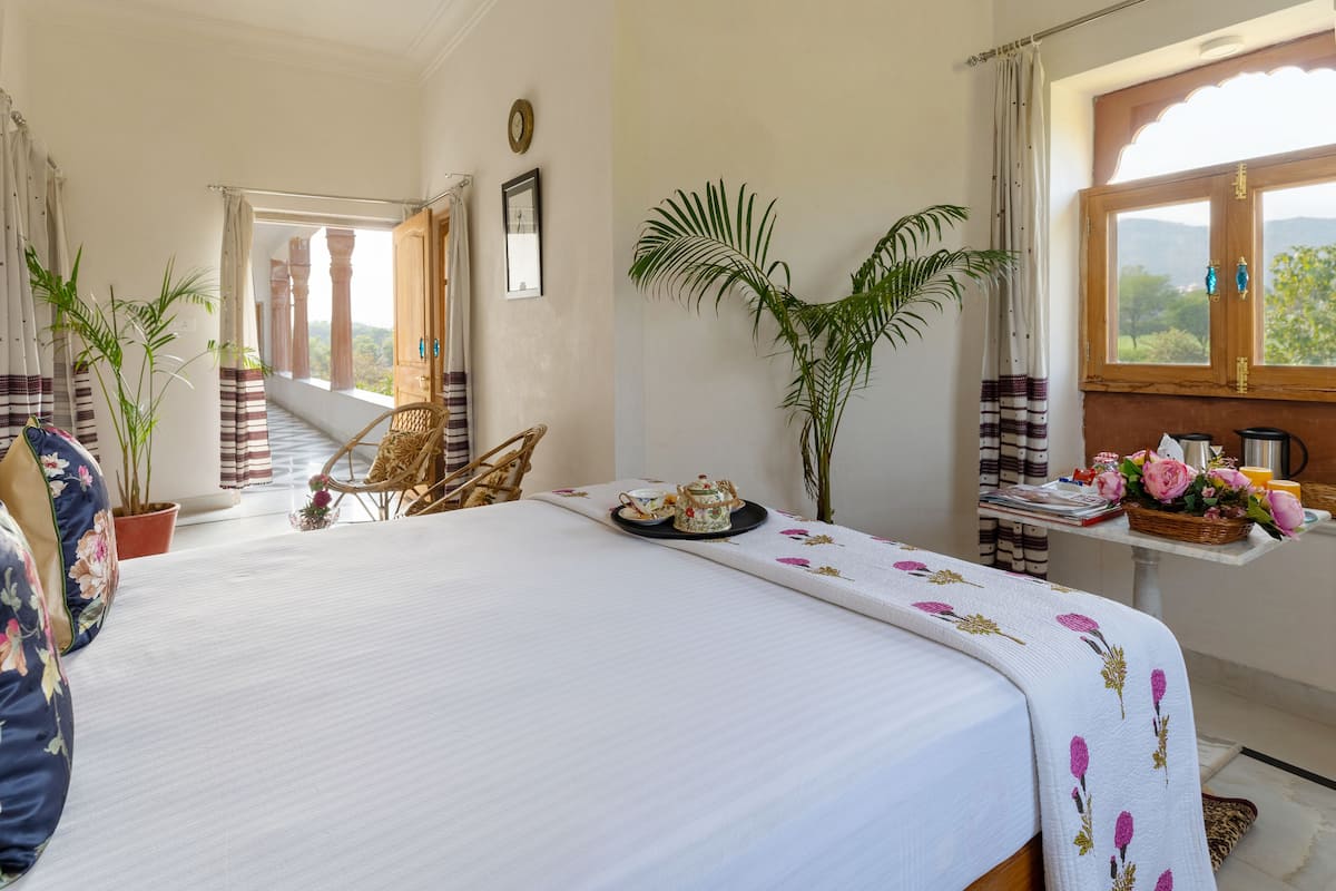 A spacious bedroom with a large bed, a wooden table with a basket of flowers, and a view of the outdoors through a window.